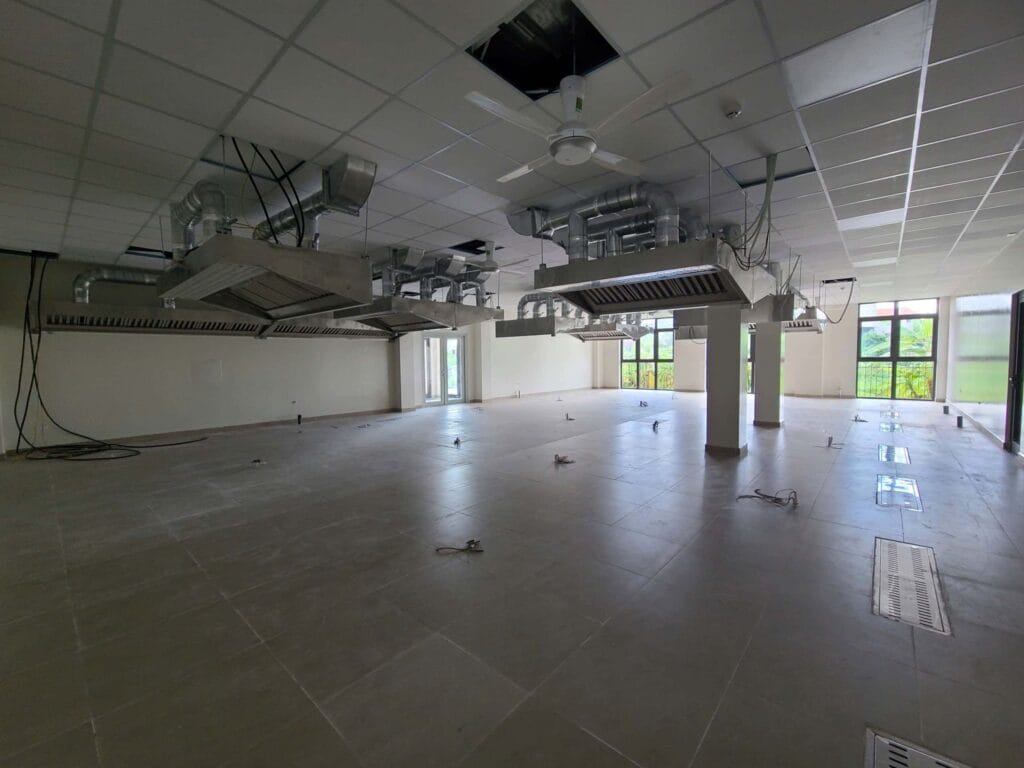 Góc nhìn toàn cảnh không gian thương mại rộng lớn đang xây dựng, nổi bật là hai hệ thống máng hút khói bếp công nghiệp bằng inox và ống thông gió đã được lắp đặt trên trần nhà lát gạch màu xám.