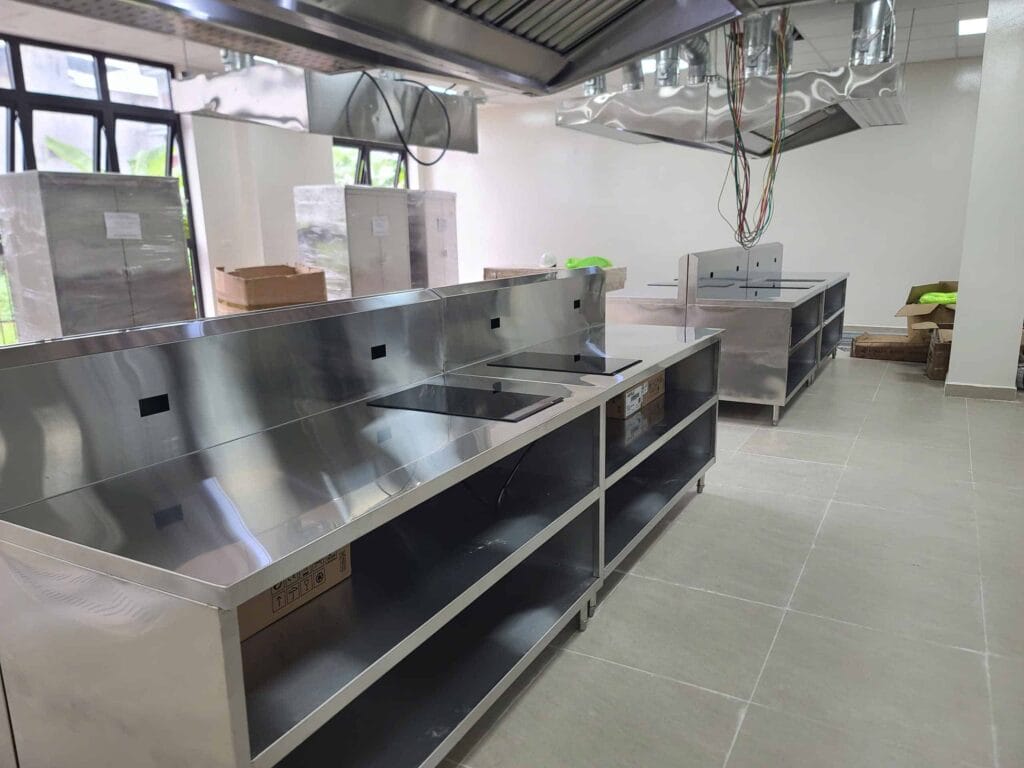 Góc chụp cận cảnh bàn bếp đảo bằng inox công nghiệp có tích hợp bếp từ, nằm dưới hệ thống hút mùi lớn, trong khu vực bếp nhà hàng/đào tạo đang trong quá trình lắp đặt thiết bị.