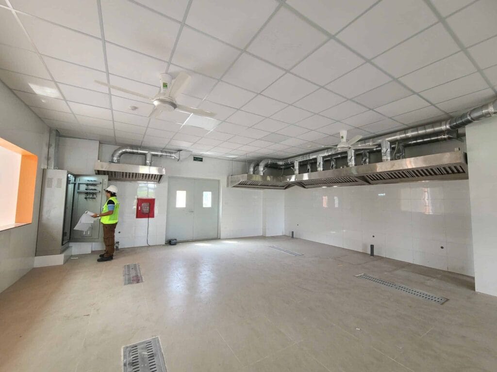 Kỹ sư kiểm tra tủ điện trong khu vực bếp công nghiệp đã ốp gạch trắng, với hai hệ thống hút mùi inox lớn và rãnh thoát nước sàn kim loại, minh họa cho dự án hoàn thiện bếp nhà hàng.