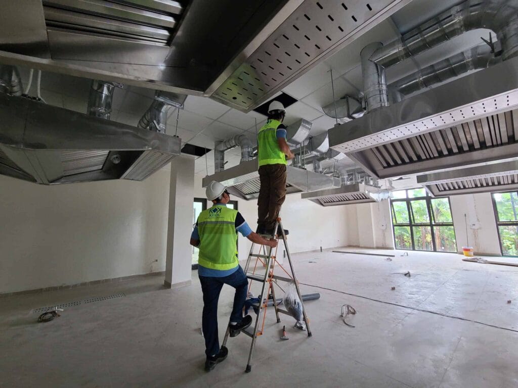 Hai công nhân đội mũ bảo hiểm và áo phản quang đang lắp đặt và kiểm tra hệ thống thông gió, máng hút mùi công nghiệp bằng thép không gỉ trên trần nhà tại công trình bếp nhà hàng đang xây dựng.